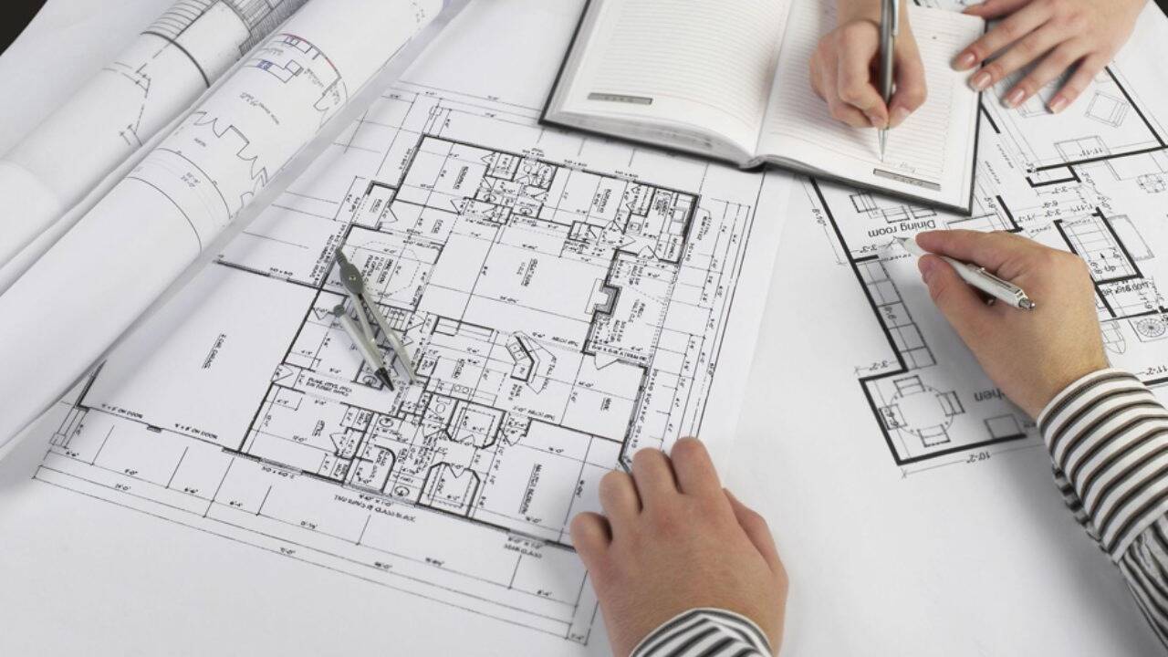 Bản vẽ thiết kế kỹ thuật nhà ở và tay người đang ghi chép kế hoạch – tượng trưng cho quá trình lập dự toán chi phí xây nhà chi tiết.
